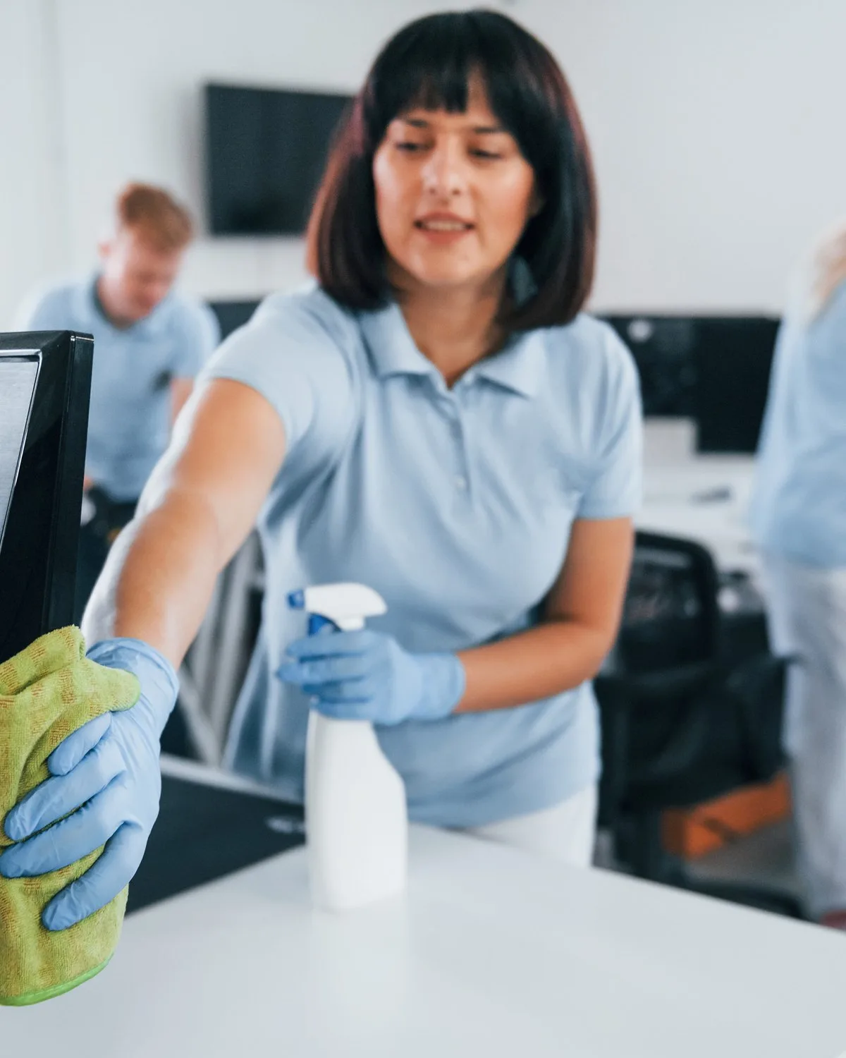 Cleaner in a light blue uniform and gloves wiping down an office workstation