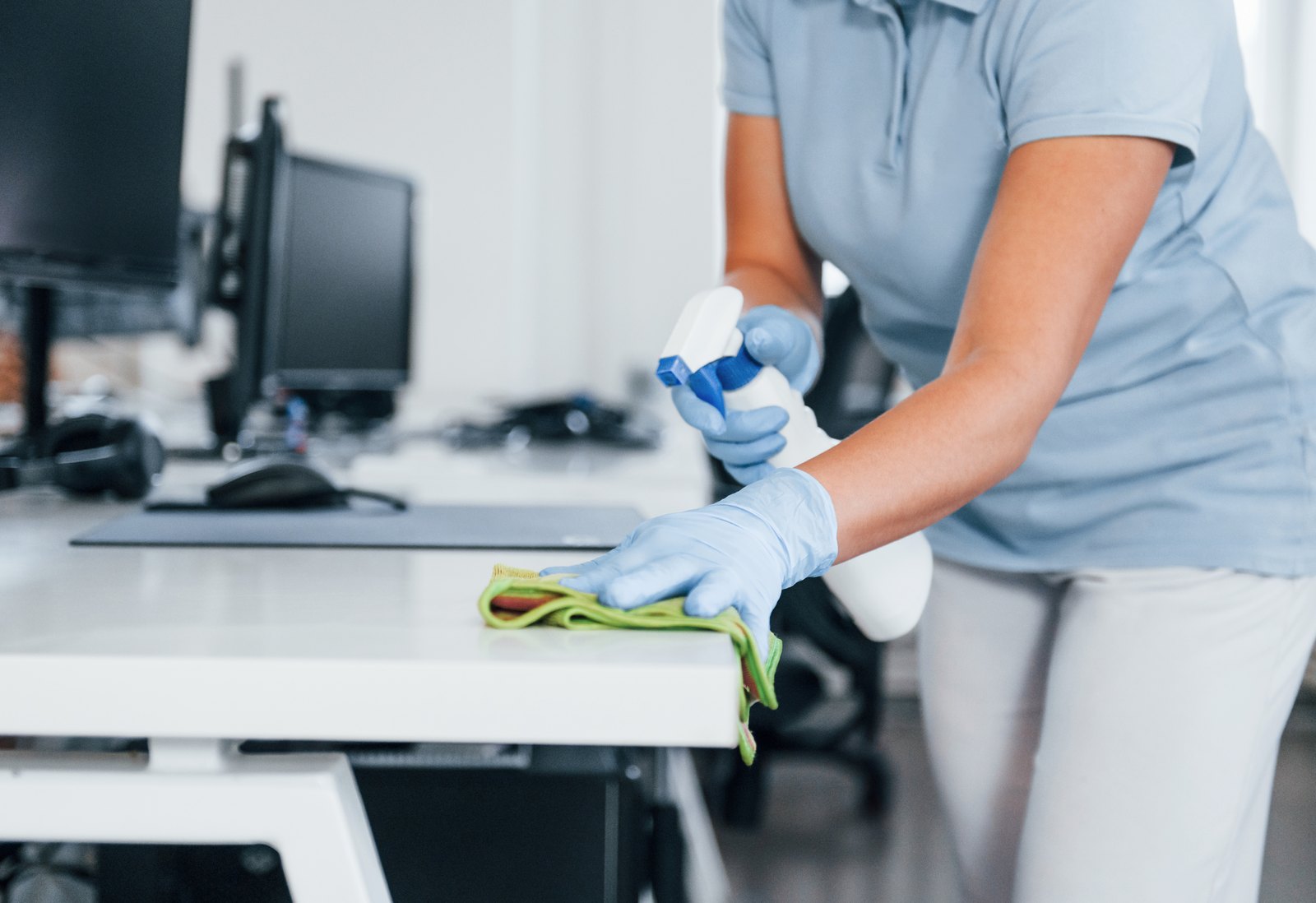 Cleaner in blue gloves wiping down a white office desk with a spray bottle and green cloth